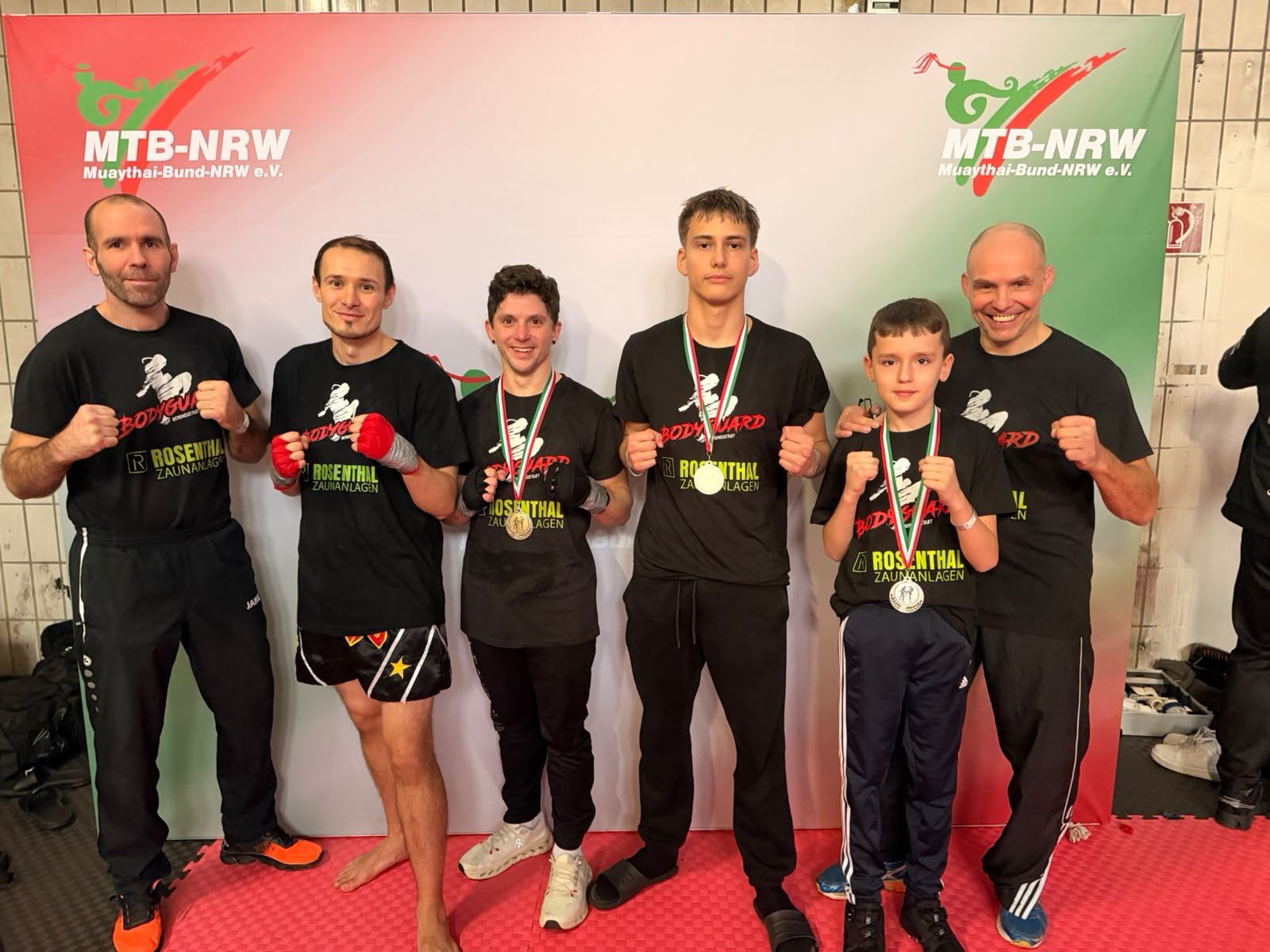 Gruppenfoto der Wettkämpfer und Trainer des Kampfsportvereins Bodyguard e.V. nach den Landesmeisterschaften 2025 des MTB-NRW in Hückelhoven. Die Sportler tragen Medaillen und stehen vor der offiziellen Fotowand des Muaythai-Bundes Nordrhein-Westfalen.